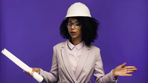 Donna afro-americana scontenta in casco di sicurezza con carta arrotolata isolata su viola — Foto stock