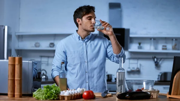 Jovem segurando vidro e bebendo vinho branco perto da mesa com ingredientes frescos — Stock Photo