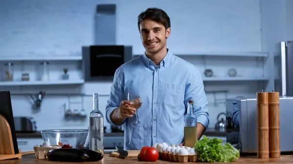 Jovem feliz segurando copo de vinho branco perto da mesa com ingredientes frescos — Fotografia de Stock