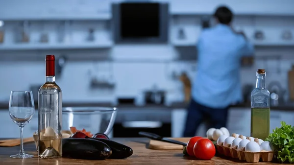 Fresh ingredients and bottle with white wine near blurred man in kitchen — Stock Photo