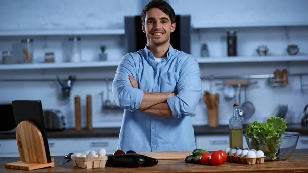 Homem sorrindo olhando para a câmera enquanto estava de pé com os braços cruzados perto da mesa com ingredientes frescos — Fotografia de Stock