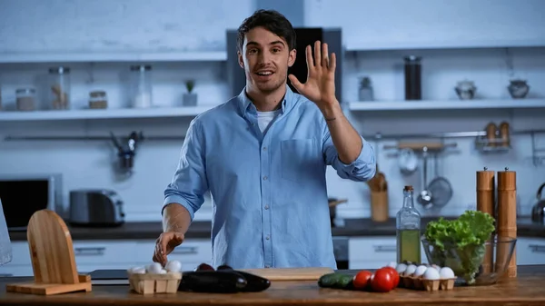 Smiling man looking at camera while waving hand near table with fresh ingredients — Stock Photo