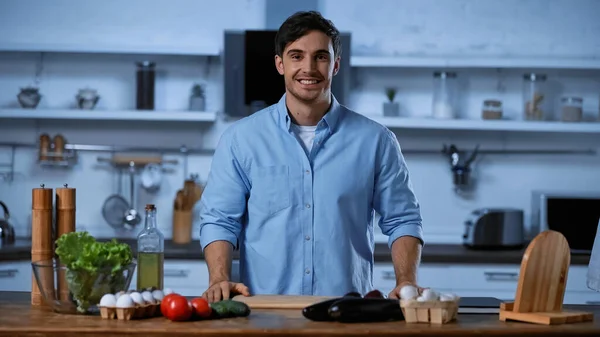 Sonriente hombre mirando a la cámara mientras está de pie cerca de la mesa con ingredientes frescos - foto de stock