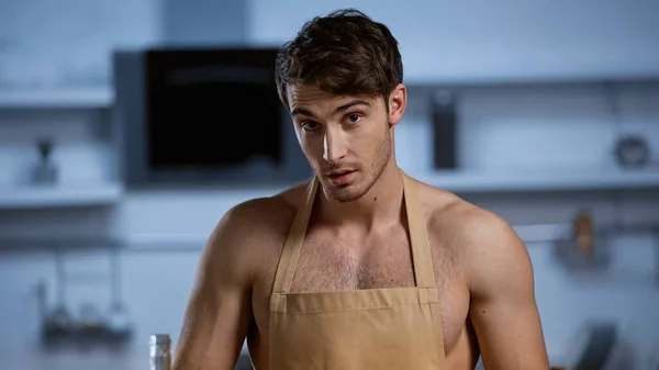 Homem sem camisa em avental bege olhando para a câmera na cozinha — Fotografia de Stock