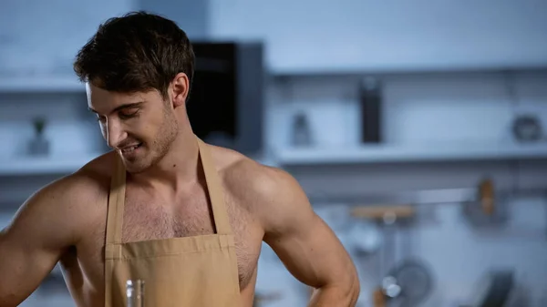 Мужчина без рубашки в фартуке улыбается и смотрит вниз на кухне — стоковое фото