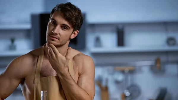 Мужчина без рубашки в фартуке смотрит в камеру, прикасаясь лицом к кухне — стоковое фото