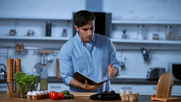 Young man reading cookbook while standing near table with fresh ingredients — Stock Photo