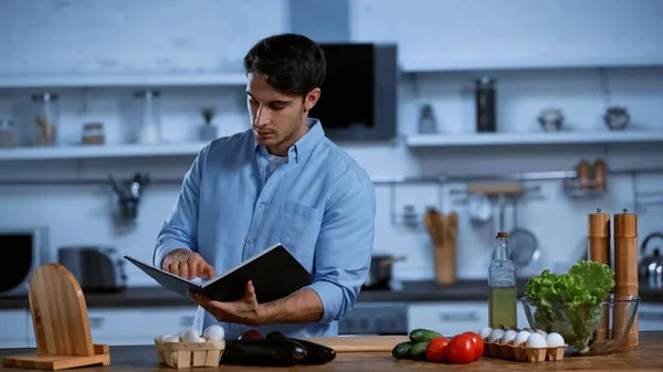 Joven leyendo libro de recetas mientras está de pie cerca de la mesa con ingredientes frescos - foto de stock