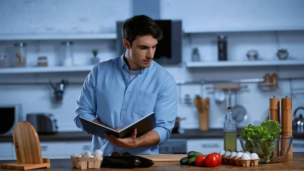 Jeune homme tenant livre de cuisine tout en regardant la table avec des ingrédients frais — Photo de stock