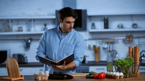 Giovane che tiene in mano il ricettario mentre guarda la tavola con ingredienti freschi — Foto stock