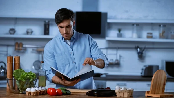 Jovem homem ler livro de receitas perto da mesa com ingredientes frescos — Fotografia de Stock