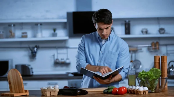 Joven hombre leyendo libro de recetas cerca de la mesa con verduras frescas - foto de stock