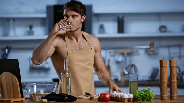 Shirtless man in apron drinking white wine in modern kitchen — Stock Photo