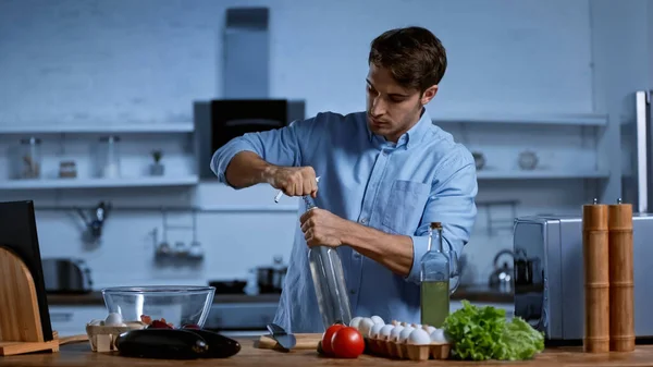 Giovane aprendo bottiglia di vino con cavatappi vicino ingredienti sul tavolo in cucina — Foto stock