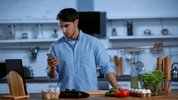 Joven sosteniendo teléfono inteligente cerca de verduras en la mesa en la cocina - foto de stock