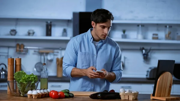 Young man holding smartphone and looking at vegetables on table in kitchen — Stock Photo