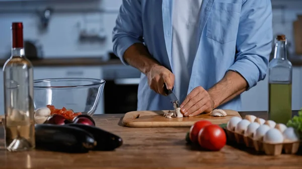 Vista ritagliata dell'uomo che taglia funghi vicino a verdure fresche, uova di pollo e bottiglia di vino — Foto stock