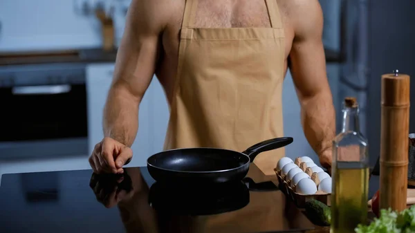Обрізаний вид без сорочки в фартусі, що стоїть біля сковороди та курячих яєць — стокове фото