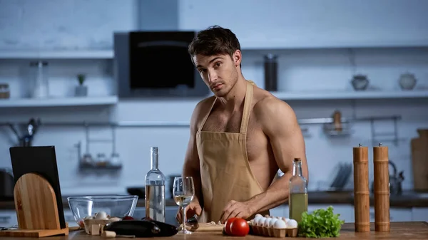 Hombre sin camisa en delantal cocina en la cocina y mirando a la cámara - foto de stock