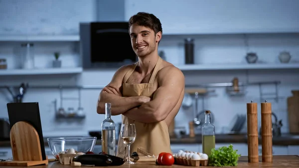 Uomo senza maglietta in grembiule posa con le braccia incrociate mentre sorride e guarda la macchina fotografica in cucina — Foto stock