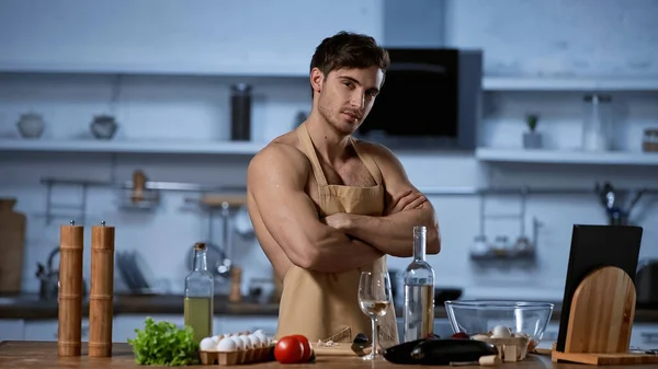 Hombre sin camisa en delantal posando con los brazos cruzados mientras mira a la cámara en la cocina - foto de stock