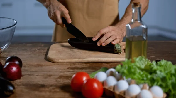 Vista parziale dell'uomo che taglia melanzane vicino a pomodori, cipolle, uova di pollo e bottiglia con olio — Foto stock