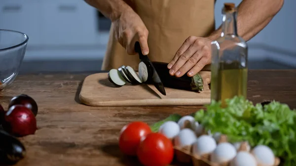 Обрезанный вид человека, режущего баклажаны возле помидоров, лука, куриных яиц и бутылки с маслом — стоковое фото