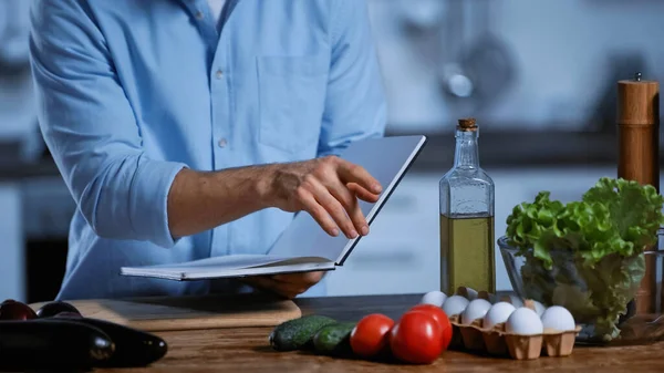 Vista recortada del hombre sosteniendo libro de cocina y apuntando a las verduras frescas en la mesa - foto de stock