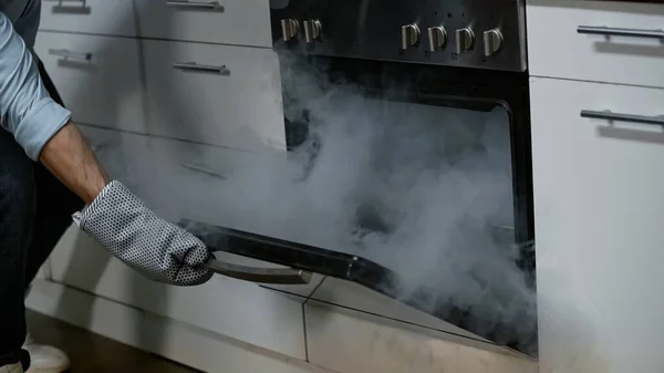 Cropped view of man in kitchen gloves opening smoking oven in kitchen — Stock Photo