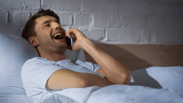 Hombre emocionado riendo mientras habla en el teléfono móvil en la cama - foto de stock