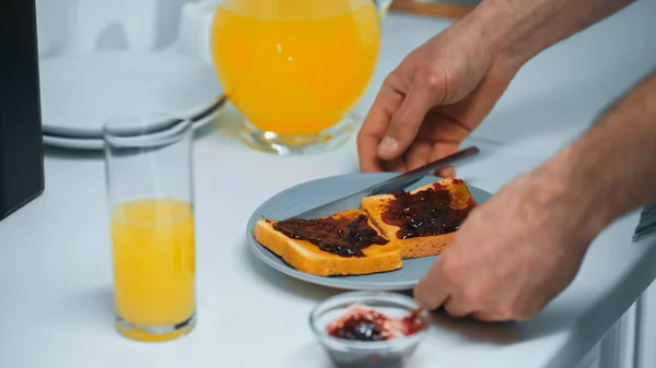 Vista parcial del hombre cerca de zumo de naranja fresco y tostadas con mermelada en la cocina - foto de stock