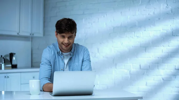 Happy freelancer in blue shirt sitting in kitchen and working on laptop — Stock Photo