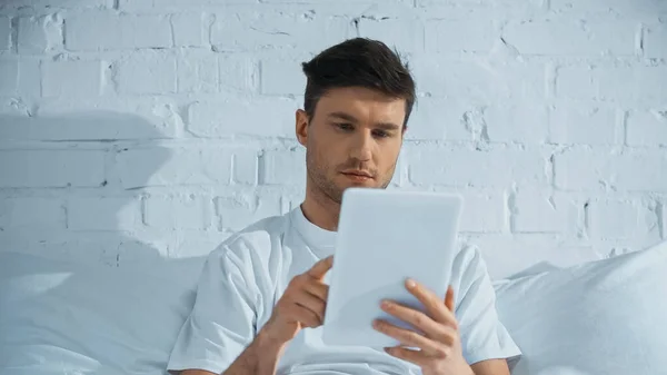 Man in white t-shirt using digital tablet while sitting on bed in morning — Stock Photo