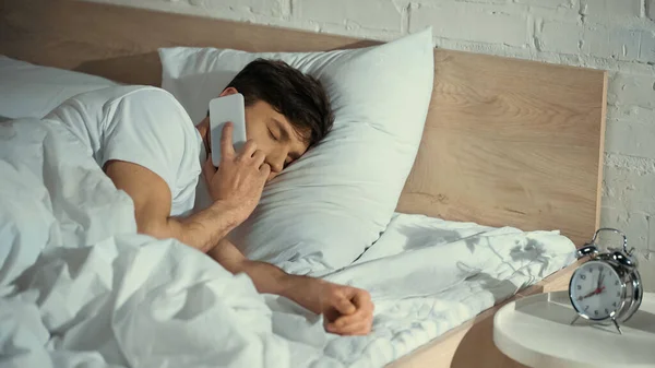 Hombre con los ojos cerrados hablando en el teléfono móvil en la cama cerca del despertador en la mesita de noche - foto de stock