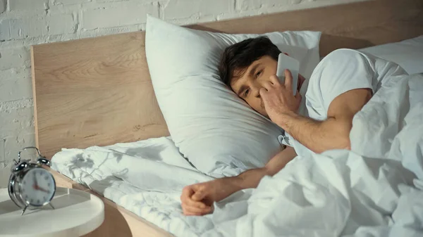 Hombre hablando en el teléfono móvil mientras está acostado en la cama cerca del reloj despertador vintage - foto de stock