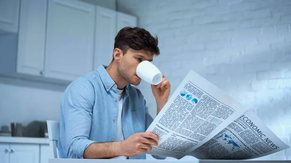 Homem de camisa azul bebendo café e lendo jornal da manhã na cozinha — Fotografia de Stock