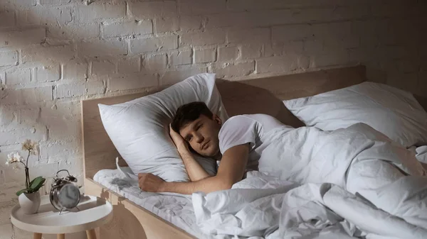 Man lying in bed near vintage alarm clock and flowerpot on bedside table — Stock Photo