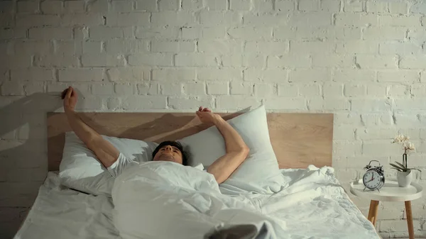 Homem esticando-se na cama perto do despertador e do orchid potted na tabela da cabeceira — Fotografia de Stock