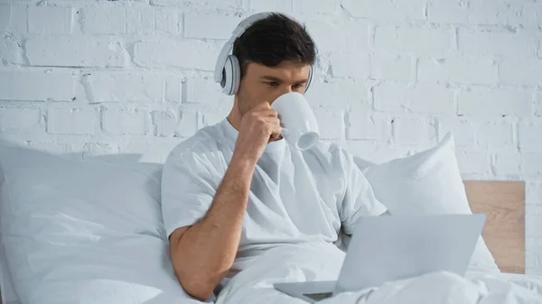 Hombre en auriculares beber café mientras se utiliza el ordenador portátil en la cama - foto de stock