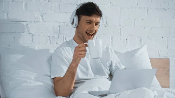 Сміється людина в навушниках тримає чашку, дивлячись на ноутбук у ліжку — стокове фото
