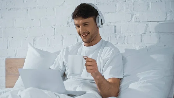 Hombre alegre en auriculares sosteniendo taza cerca del ordenador portátil en la cama - foto de stock