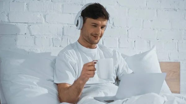 Усміхнений чоловік в навушниках тримає чашку, використовуючи ноутбук у ліжку — стокове фото