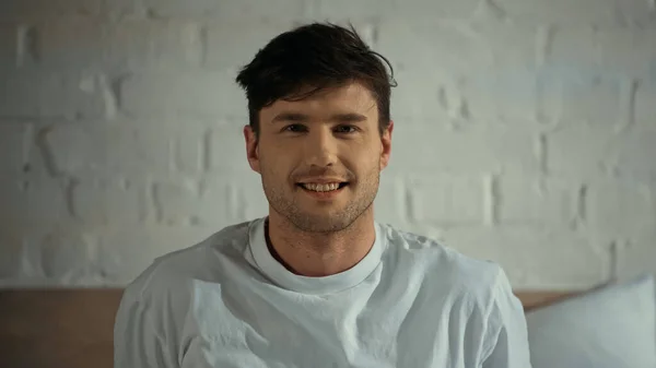 Hombre en camiseta blanca sonriendo a la cámara en el dormitorio - foto de stock