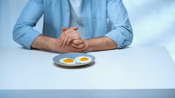 Обрезанный вид человека, сидящего с сжатыми руками возле тарелки с жареными яйцами — стоковое фото