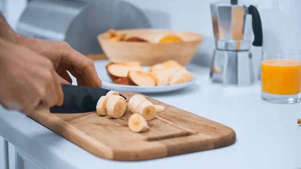 Vista recortada del hombre cortando plátano fresco en la tabla de cortar en la cocina - foto de stock