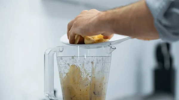 Vista cortada do homem adicionando banana cortada em tigela de liquidificador enquanto prepara o smoothie — Fotografia de Stock