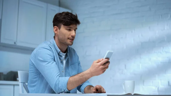 Улыбающийся мужчина болтает по мобильному телефону, сидя на кухне — стоковое фото