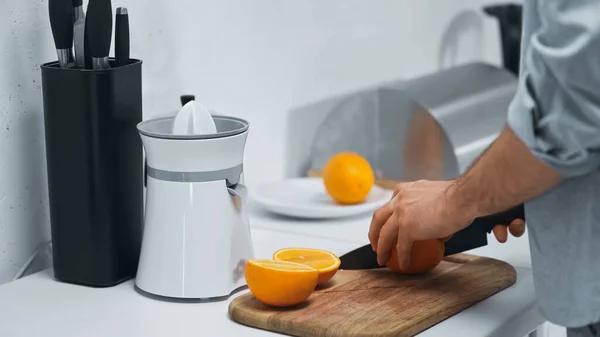 Vista parcial del hombre cortando naranja fresca cerca de exprimidor eléctrico en la cocina - foto de stock