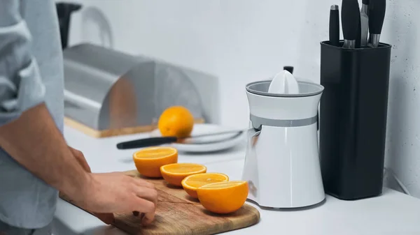 Vista recortada del hombre cerca de mitades de naranjas frescas y exprimidor eléctrico en la cocina - foto de stock