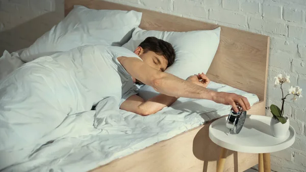 Людина з закритими очима лежить в ліжку і вимикає будильник — стокове фото